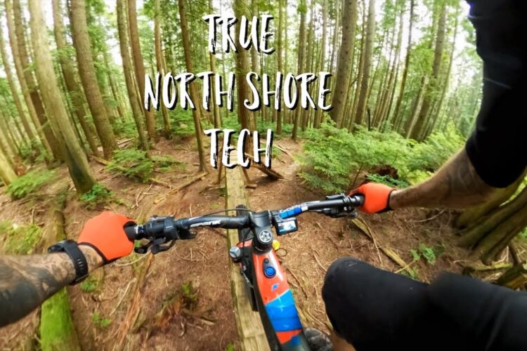 A mountain biker's perspective while navigating a narrow wooden bridge through a dense forest, with vibrant greenery and tall trees surrounding the trail. The biker's hands are gripping the handlebars adorned with bright orange gloves, showcasing a close-up view of the bike's controls. The text "TRUE NORTH SHORE TECH" is overlaid at the top of the image.