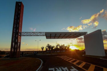 A modern architectural entrance structure with a flat canopy and a tall vertical element, set against a vibrant sunset sky. Silhouettes of palm trees are visible in the background, and a winding road leads towards the entrance. The foreground features road markings indicating a parking area. Trilha do parque da cidade mountain bike trail.