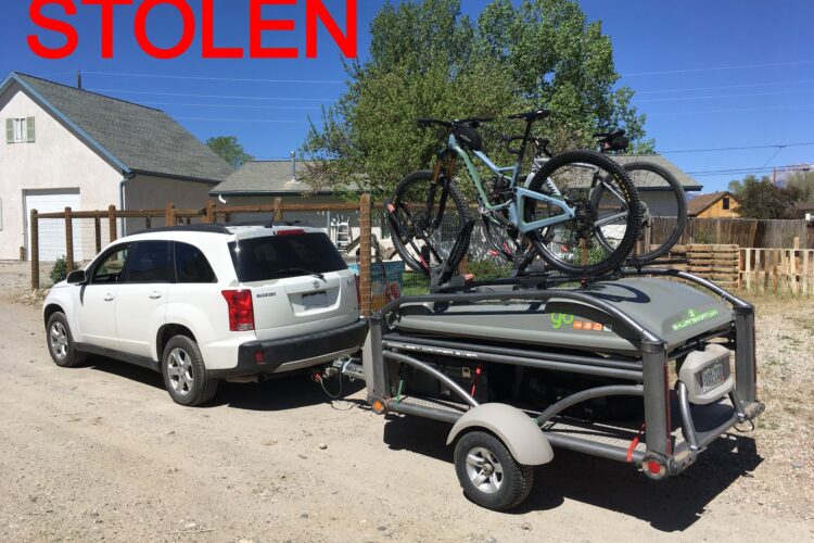 A white SUV parked on a dirt road, towing a small trailer with two bicycles mounted on top. The trailer has a logo on it and is labeled with the word "STOLEN" in large red letters. In the background, there are trees and a house visible.