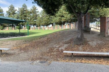 A paved area leading to a grassy hill, with a shaded picnic structure and benches. Lush green trees are visible in the background, and the ground is scattered with fallen leaves. Grass Peak Trail mountain bike trail.
