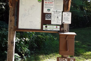 A wooden information board at the Endless Brook Trails, displaying a map of the area along with various trail guidelines and alerts for visitors. A brown sign-in box is positioned at the base, and a "Sign In" sign hangs nearby. The surrounding area features greenery typical of a forest setting. Endless Brook Trails mountain bike trail.