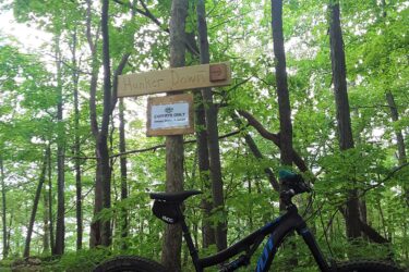 A mountain bike is leaning against a tree in a forested area. In the background, a wooden sign labeled "Hunker Down" is visible, pointing to the right. Below it, there is a sign that reads "EXPERTS ONLY." The scene is surrounded by lush green foliage, indicating a vibrant outdoor setting. Endless Brook Trails mountain bike trail.