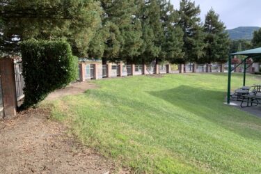 A grassy area with a slope, surrounded by tall trees and a brick wall with white railings. A neatly trimmed hedge is visible on the left, and a shaded picnic area with tables is located on the right. In the background, a mountain range is faintly visible under a cloudy sky. Grass Peak Trail mountain bike trail.