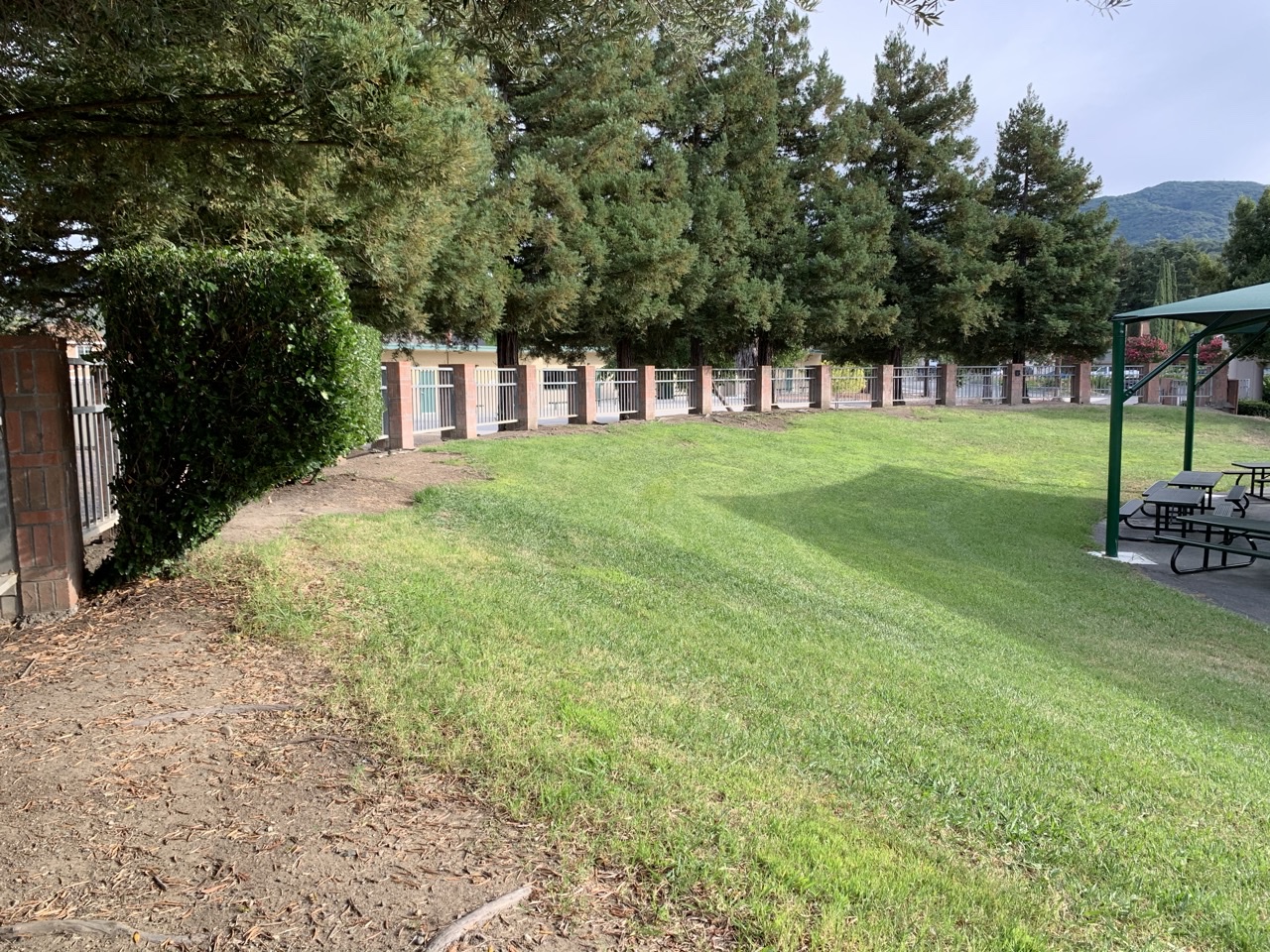 A grassy area with a slope, surrounded by tall trees and a brick wall with white railings. A neatly trimmed hedge is visible on the left, and a shaded picnic area with tables is located on the right. In the background, a mountain range is faintly visible under a cloudy sky. Grass Peak Trail mountain bike trail.