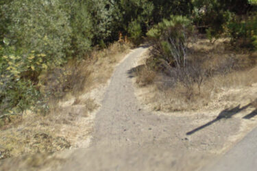 A narrow dirt path winding through a dry landscape, bordered by sparse vegetation and shrubs, with a nearby wooden post casting a shadow on the ground. Alamitos Creek Trail mountain bike trail.