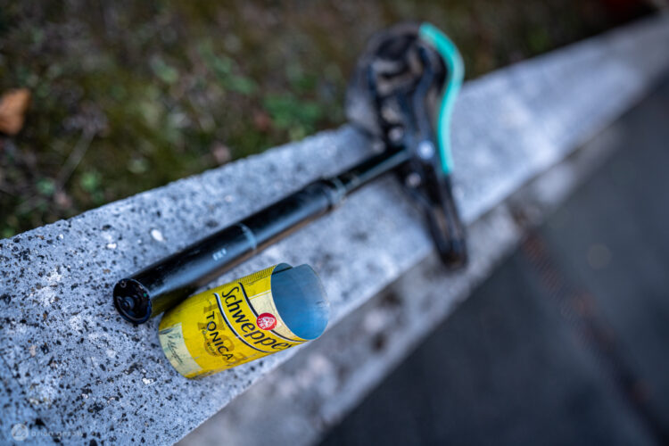 A black handle with a turquoise accent rests on a concrete ledge next to a partially opened can of Schweppes tonic water. The background shows blurred greenery, hinting at an outdoor setting.
