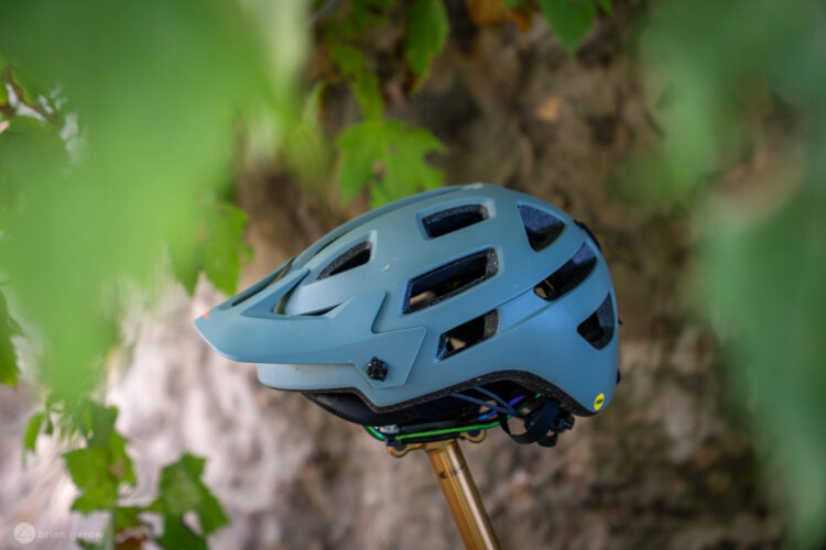 A side view of a light blue bicycle helmet with ventilation holes, resting against a textured rock surface and partially obscured by green leaves. The helmet features a visor and adjustable straps.