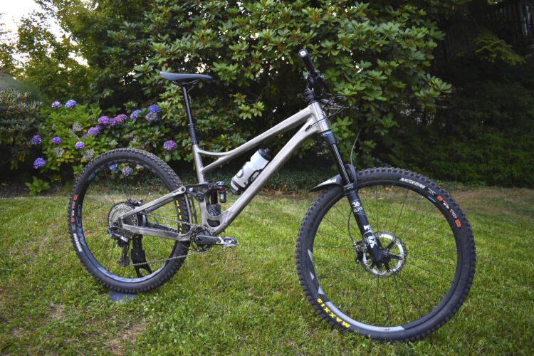A silver mountain bike with thick tires and a water bottle holder stands on green grass, surrounded by flowering bushes in the background. The bike features front suspension, a geared rear system, and a sleek frame design.