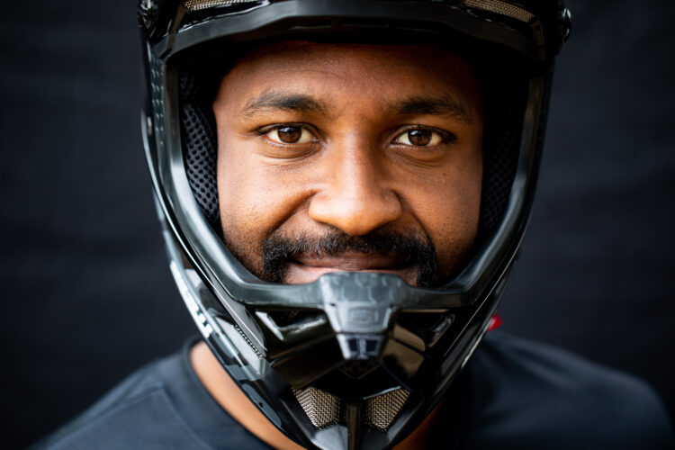 Close-up portrait of a man wearing a black motorcycle helmet, smiling gently at the camera, with a neutral dark background. He has a short beard and brown eyes, and is dressed in a black shirt.