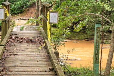 A wooden bridge with caution tape and signs on either side crosses a river with muddy, brown water. The bridge, labeled "Advance America Bridge," is surrounded by lush greenery and trees. Debris is scattered across the bridge, indicating recent flooding or storm activity. Lake Johnson Fairforest Creek Connector mountain bike trail.