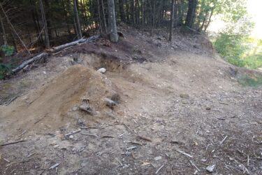 A dirt area with uneven mounds and a cleared space, surrounded by trees and scattered twigs. The ground is mostly bare with patches of soil and rocks visible. Pineland Trail mountain bike trail.