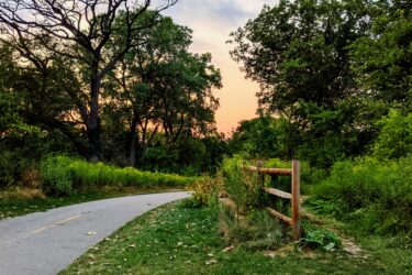 A winding pathway through a lush green landscape, bordered by trees and a wooden fence, under a colorful sky at dusk. The scene conveys a tranquil and natural outdoor setting. Forest Glen Woods mountain bike trail.