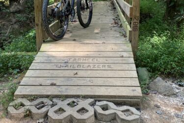 A mountain bike is leaning against a wooden bridge in a wooded area. The bridge features engraved text reading "Tarheel Trailblazers." In front of the bridge, there are decorative concrete blocks arranged on the ground, surrounded by natural foliage and dirt. The scene is lush and green, showcasing a peaceful outdoor setting. Colonel Francis Beatty Park mountain bike trail.