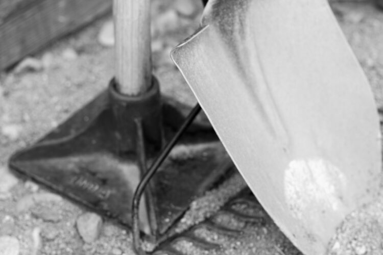 A close-up black and white image of a metal shovel resting on gravel, showcasing the shovel's blade, handle, and base. The background features a textured surface with scattered pebbles.