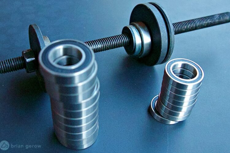 A close-up image of metallic ball bearings arranged in stacks, with a threaded rod and a flat disc component in the background. The bearings are shiny and cylindrical, showcasing their smooth surfaces against a dark background.