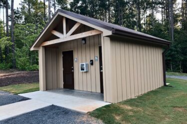 A small, newly constructed restroom building with a peaked roof, made of wood and metal siding. Two entrance doors are visible, each labeled with gender symbols. A payment kiosk is mounted on the wall, and the building is surrounded by greenery and trees in the background. A concrete path leads up to the entrance, with gravel landscaping nearby. Brown's Mill mountain bike trail.