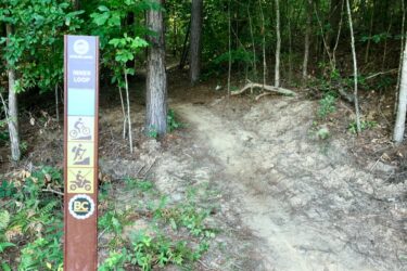 A sign marking the entrance to the "Inner Loop" trail in a wooded area, featuring icons for biking, hiking, and off-road motorcycling, surrounded by lush greenery. Brown's Mill mountain bike trail.
