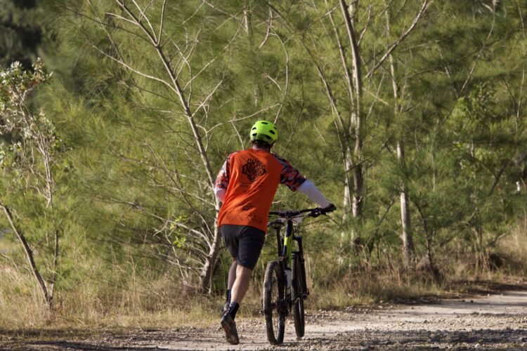 A person wearing an orange shirt and a yellow helmet is walking their mountain bike along a gravel path surrounded by trees. The scene is set in a natural outdoor environment with greenery in the background.