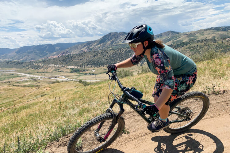 A person riding a mountain bike on a dirt trail, surrounded by a scenic landscape of hills and valleys under a partly cloudy sky. The cyclist is wearing a helmet and sunglasses, and is dressed in a floral-patterned outfit. The bike is in motion, highlighting the action of mountain biking.