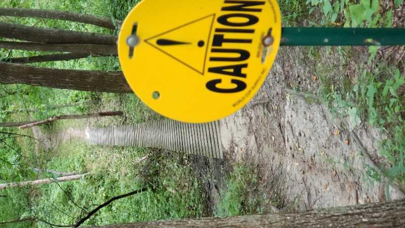Caution sign in a wooded area indicating a warning, with a wooden pathway visible in the background among trees and foliage. Beverly Park mountain bike trail.