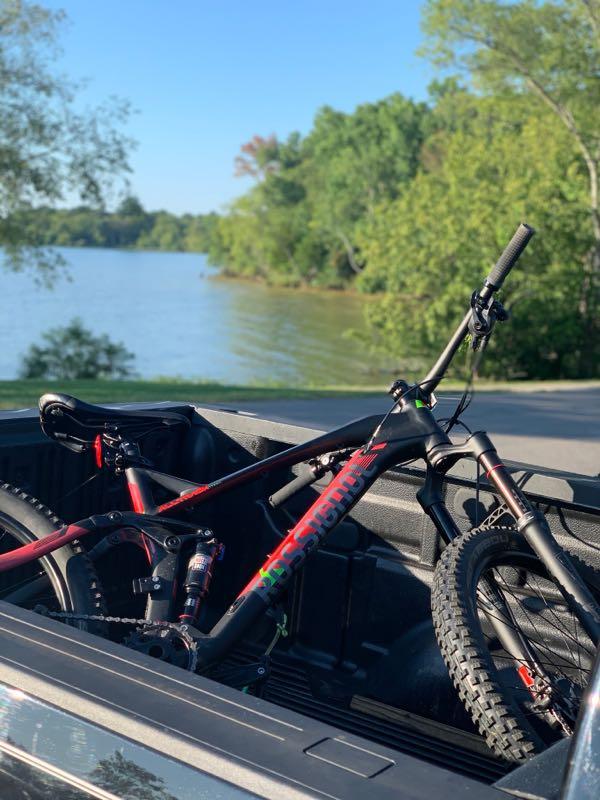 A mountain bike rests in the bed of a pickup truck, with a scenic lake and green trees in the background. The image captures a bright sunny day, showcasing the bike’s vibrant colors against the serene natural setting. Lock 4 mountain bike trail.