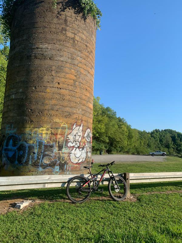 A mountain bike rests beside a large, weathered silo covered in colorful graffiti. The background features a clear blue sky and lush green trees, while a gravel parking area is visible nearby with a vehicle parked in the distance. Lock 4 mountain bike trail.