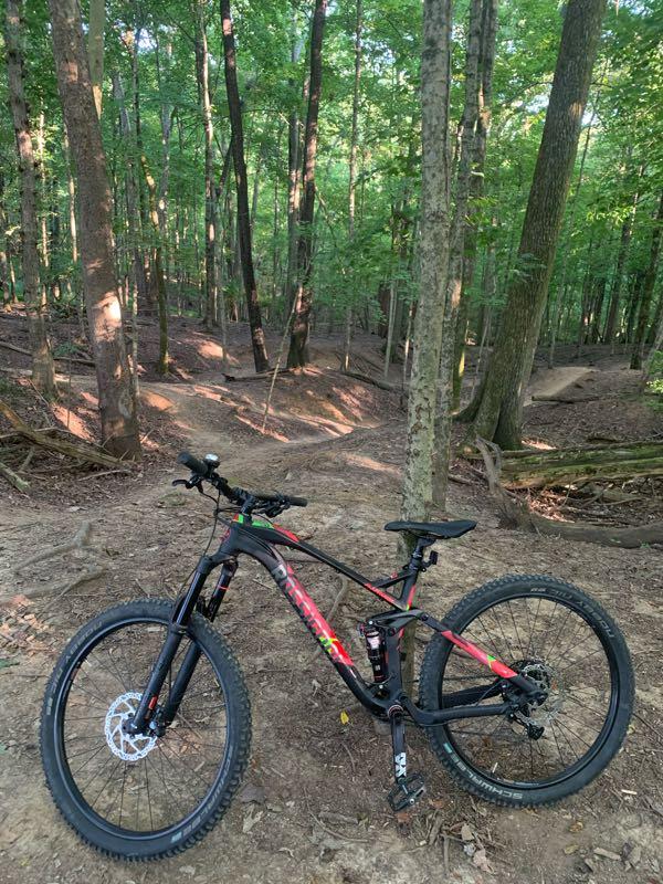 A mountain bike parked on a dirt trail surrounded by dense green trees in a forested area. The path winds through the woods, featuring sunlight filtering through the foliage. Lock 4 mountain bike trail.