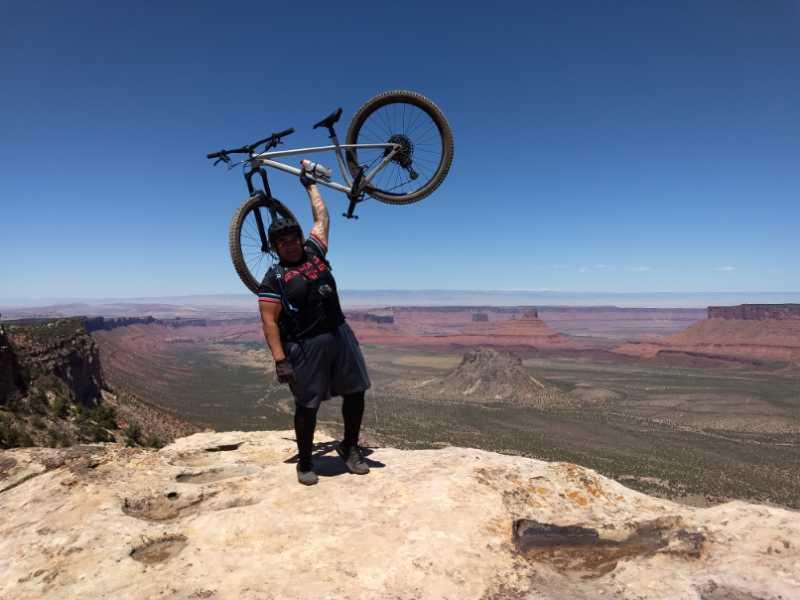 A mountain biker stands triumphantly on a rocky outcrop, raising his bicycle above his head against a backdrop of vast, rugged landscapes and blue skies. The scene captures a sense of achievement and adventure in an outdoor setting.