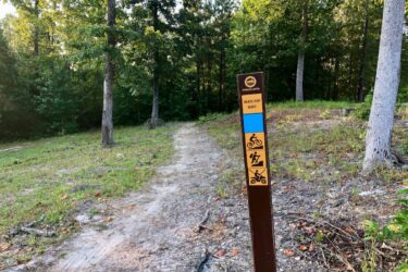 Trail sign indicating the "Black Dirt Bluff" biking trail, featuring symbols for biking, hiking, and off-road vehicles, surrounded by a wooded area with trees and a dirt path. Moore's Bridge mountain bike trail.