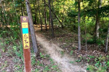Signpost marking the entrance to the Black Dirt Bluff trail in a forested area, with a dirt path leading into the woods. The sign indicates bike riding, hiking, and off-road vehicle use. Lush greenery surrounds the trail, with trees and underbrush visible. Moore's Bridge mountain bike trail.