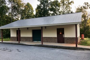 One-story building with a metal roof, featuring two dark doors and a green roller shutter. The lower section of the walls is made of stone, and there are two light fixtures on the front. The building is surrounded by trees, with a road and a marked construction barrel visible in the foreground. Moore's Bridge mountain bike trail.