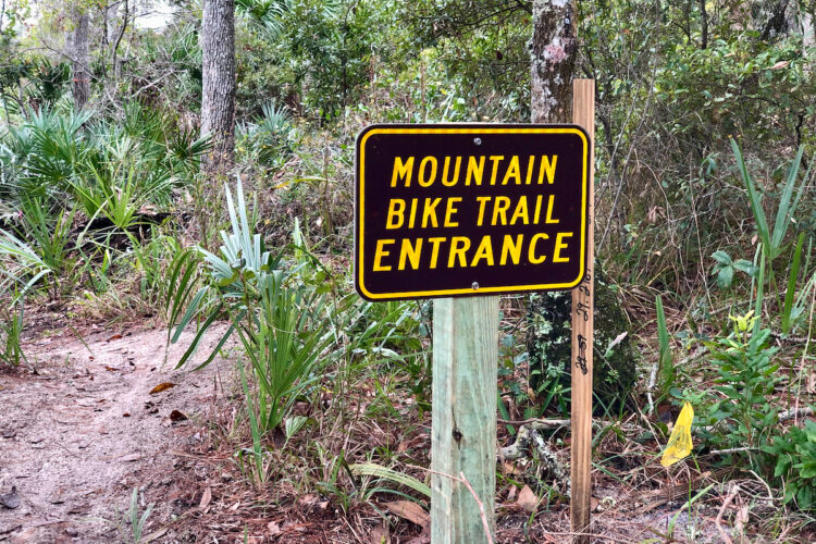 A sign marking the entrance to a mountain bike trail, set amidst a forested area with various plants and trees. The sign features bold yellow text on a dark background, indicating "MOUNTAIN BIKE TRAIL ENTRANCE."