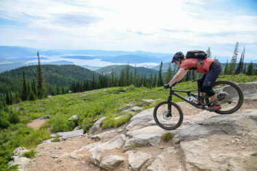 A mountain biker navigating rocky terrain on a downhill trail, surrounded by lush greenery and distant hills, with a serene lake visible in the background under a partly cloudy sky. Schweitzer Mountain Resort mountain bike trail.