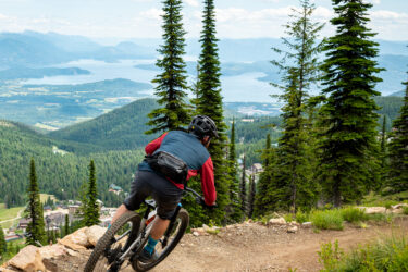 A mountain biker descends a winding trail through lush green trees with a stunning view of a lake and mountains in the background. The rider is focused on navigating the path, showcasing a sense of adventure in a scenic landscape. Schweitzer Mountain Resort mountain bike trail.