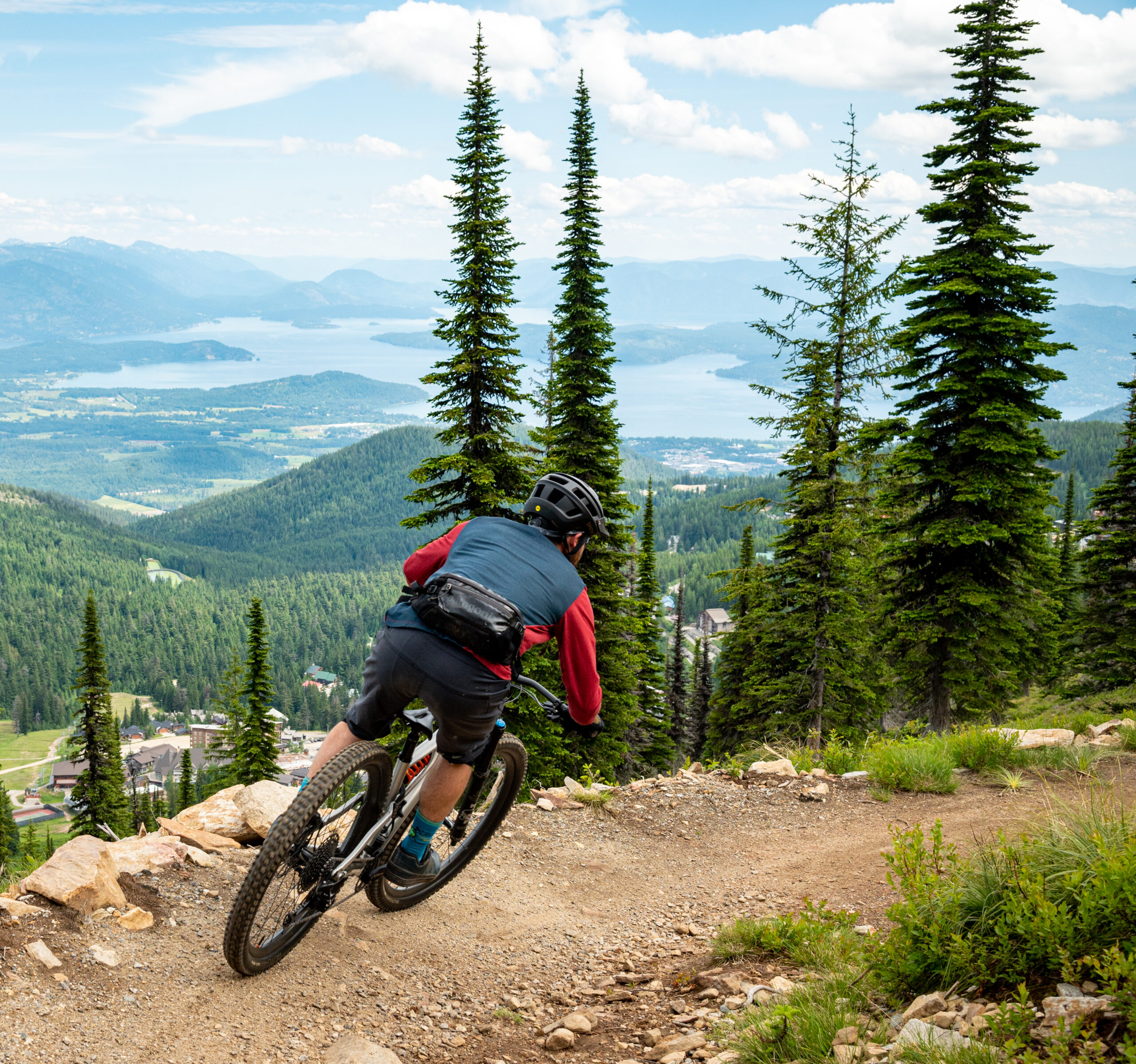 A mountain biker descends a winding trail through lush green trees with a stunning view of a lake and mountains in the background. The rider is focused on navigating the path, showcasing a sense of adventure in a scenic landscape. Schweitzer Mountain Resort mountain bike trail.