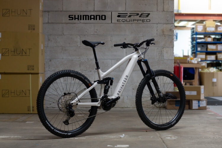 A white electric mountain bike with a sleek design, featuring a Shimano EP8 motor, standing in a warehouse setting with cardboard boxes in the background. The bike has rugged tires suitable for off-road terrain and displays the brand name "Privater" on its frame.