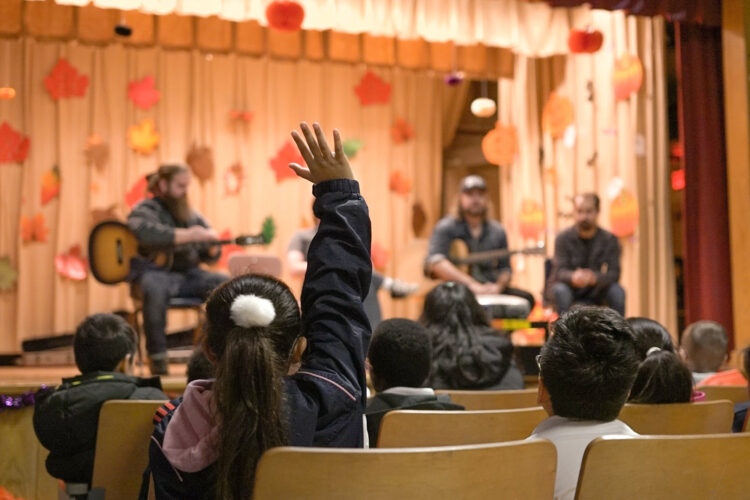 A child with a raised hand sits in an audience, eagerly participating in a music performance on stage. Several musicians are seated in front of a decorated background, featuring colorful autumn leaves. The atmosphere is lively and engaging, with other children watching attentively.