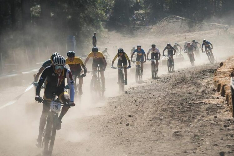 A group of mountain bikers racing on a dusty trail, surrounded by trees and a few spectators in the background. The riders are wearing colorful jerseys and helmets, showcasing determination as they maneuver through the course. Dust is kicked up around them, highlighting the intensity of the competition.