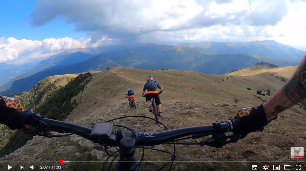 Three mountain bikers ride down a rocky trail on a hillside, with expansive green and blue mountainous scenery in the background. The foreground shows the handlebars of the bike in focus, emphasizing the thrilling descent. The sky is partly cloudy, adding to the dynamic outdoor atmosphere. Spain e-bike Singletracks mountain bike trail.