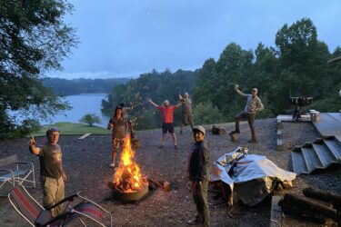 A group of six people gathered around a large campfire near a lake during twilight. They appear to be enjoying a fun outdoor gathering, with five individuals striking playful poses and one person holding a drink. Several chairs are set up nearby, and there are trees and a hillside in the background. Cub Creek Trails mountain bike trail.