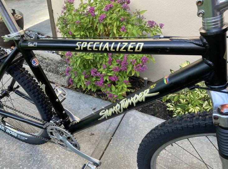 Specialized Specialized Stumpjumper A1 FS Mountain Bike: Close-up view of a black Specialized Stumpjumper bicycle frame, featuring bold yellow lettering. The bike is set against a backdrop of vibrant green and purple foliage, with visible textured tires and a shiny silver pedal. The frame displays branding and details characteristic of a mountain bike.