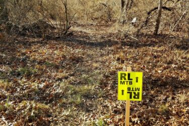 A dirt pathway surrounded by bare trees and brush, with a bright yellow sign indicating "RL MTB" pointing in two directions. The ground is covered with fallen leaves, suggesting an outdoor trail suitable for mountain biking. Rend Lake Mountain Bike Trail mountain bike trail.