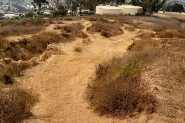 A winding dirt path surrounded by dry grass and shrubs, leading through a hilly landscape with a distant view of a city and overcast sky in the background. Sanjon Barranca mountain bike trail.