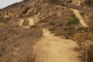 A dirt trail winding through dry, hilly terrain with sparse vegetation and shrubs. The path leads up a slope, surrounded by a muted landscape under a cloudy sky. Sanjon Barranca mountain bike trail.