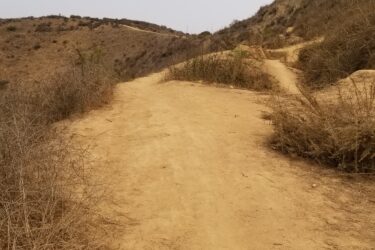 A dry, sandy hiking trail winding through a rugged landscape, surrounded by sparse vegetation and hills in the background under a clear sky. Sanjon Barranca mountain bike trail.