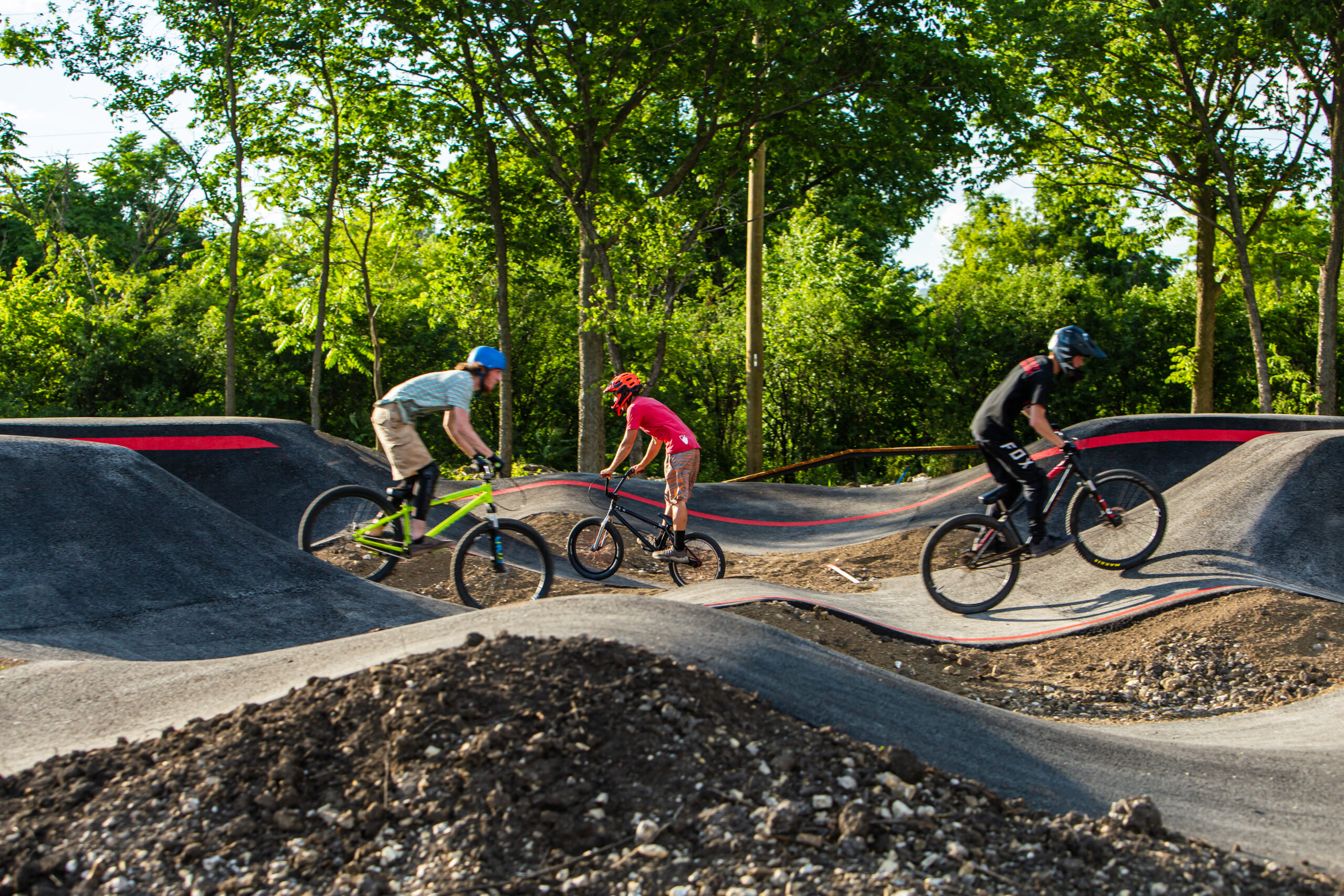 Three riders on bicycles navigate a dirt pump track surrounded by trees. The track features a series of raised, undulating bumps with red accents. The riders are wearing helmets and riding different styles of bikes, showcasing an active outdoor scene. The Forge: Lemont Quarries mountain bike trail.