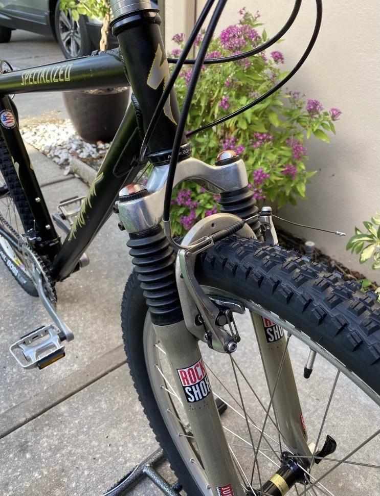 Specialized Specialized Stumpjumper A1 FS Mountain Bike: A close-up view of the front of a mountain bike, showcasing the suspension fork, the tire, and part of the bike frame with a "Specialized" logo. Surrounding foliage and a gravel pathway are visible in the background.