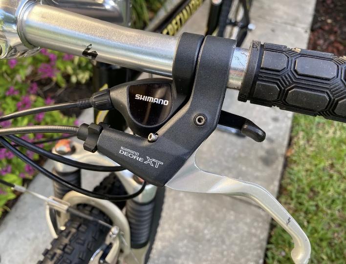 Specialized Specialized Stumpjumper A1 FS Mountain Bike: Close-up of a bicycle's handlebar featuring a Shimano Deore XT brake lever, with black rubber grips and attached cables, set against a backdrop of greenery and flowers.