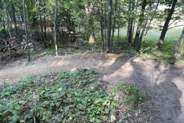 A dirt bike trail in a forested area featuring a smooth jump ramp, surrounded by trees and overgrown vegetation, with sunlight filtering through the leaves. Standing Rocks mountain bike trail.