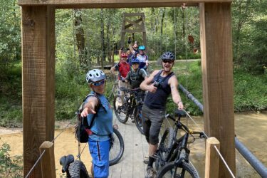 A group of enthusiastic mountain bikers poses on a wooden bridge surrounded by lush greenery. They are wearing helmets and outdoor gear, with some holding their bikes while others strike playful poses. The scene captures a sense of camaraderie and adventure in a natural setting. Cub Creek Trails mountain bike trail.